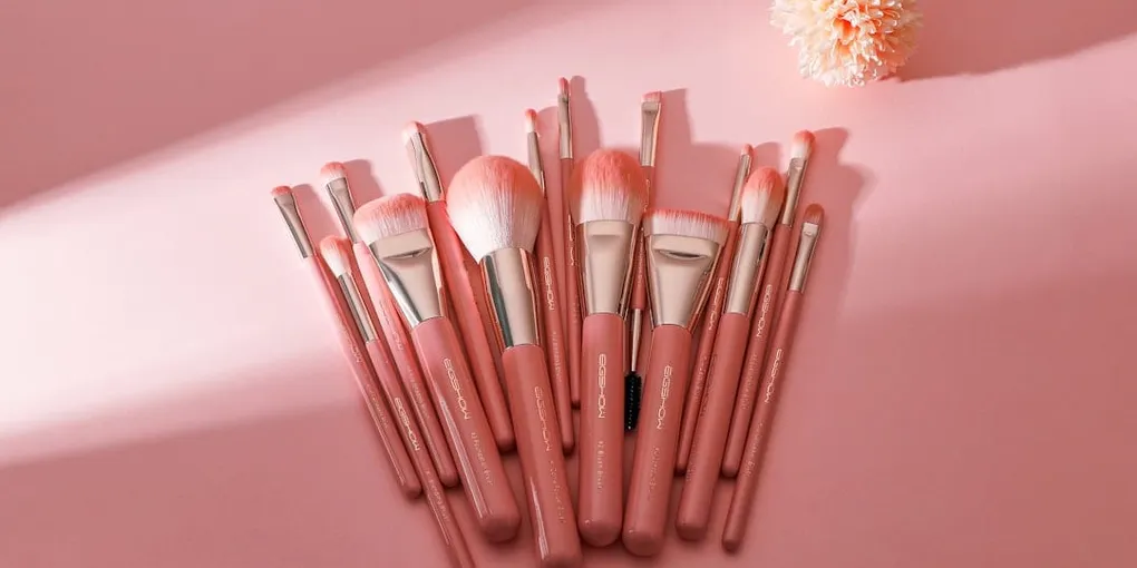 A set of pink-handled makeup brushes fanned out on a pink surface next to a dried flower.