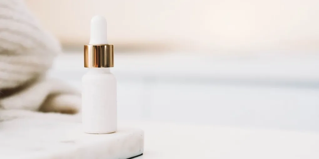 A white dropper serum bottle with a gold cap standing on a marble surface
