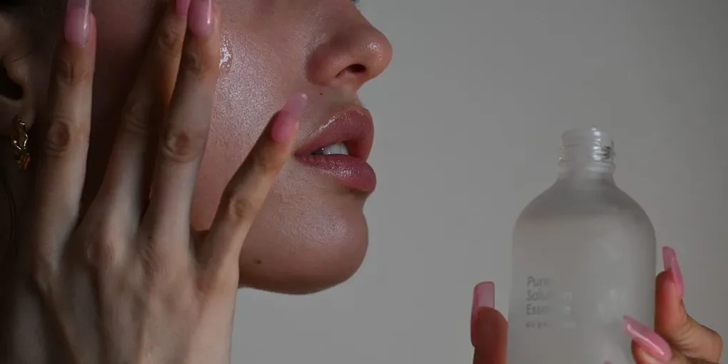 A person applying skincare toner to the face while holding a glass toner bottle.